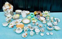 Photo shows a spread of assorted miniature fine bone china and porcelain tea set pieces arranged on a table, including cups, saucers, plates, teapots, creamers, and decorative jars in various colors and patterns.