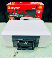 Photo of the closed white Sentry 1100 Fire-Safe security chest with black handle and lock, two keys placed in front, original product box in the background