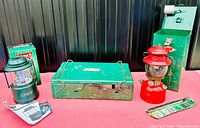 View of the entire Coleman outdoor bundle showing the green pack-away lantern, green stove with metal case, red vintage lantern, and accessories on a red surface.