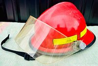 Side angle of vintage red firefighter helmet showing the clear visor, yellow reflective band, and black adjustable chin strap.