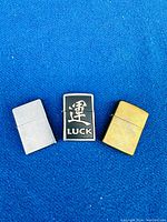 Front view of three Zippo lighters showing brass, brushed chrome, and luck symbol designs on blue background.