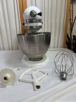 KitchenAid mixer with stainless steel bowl and three attachments (dough hook, flat beater, wire whisk) arranged in front of it on white cloth.