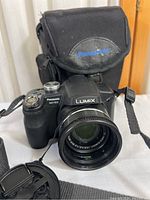 Front view of Panasonic Lumix DMC-FZ18 camera with attached Leica lens, lens cap, and Panasonic branded carrying case behind camera.