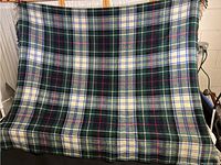 Full view of wool tartan blanket in green, blue, cream, and red with fringed edge spread out on chair or rack.