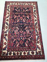 Full view of the hand knotted wool Persian rug showing the central dark colored field with intricate floral patterns and the cream border with red and blue accents.