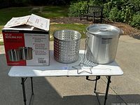 Full set of the boiling pot with the box, perforated basket, lid and star-shaped pot holder stand all shown on a table outside in daylight.
