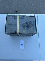 Top view of the original box showing the product name and illustration of the bench vise, box secured with a yellow strap.