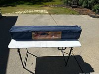 Blue 9x9 pop up gazebo canopy folded inside a carry bag placed on a white table on concrete surface outdoors.