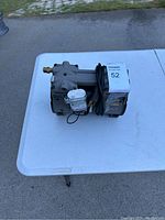 Metal AC vacuum pump placed on white outdoor table, showing attached motor and electrical wiring.