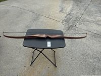Full view of the wooden traditional recurve bow laid out flat on a folding table outdoors on concrete surface.