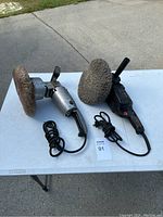 Two electric car buffers with round buffing pads and power cords placed on table next to auction lot tag