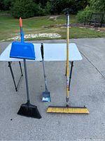 Photo showing two brooms and one metal rake scoop set on a white folding table outside on concrete surface.