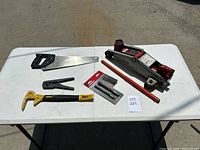 Full view of all tools on white table including car jack, saw, hammer, pry bar, wire stripper, drill bits, and extension rod