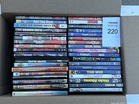 Top-down view of a cardboard box filled with 75 assorted DVDs showing titles such as "Head of State," "Paint Your Wagon," "Are We Done Yet?", "Two Can Play That Game", "The Pursuit of Happyness", "Heart and Souls", "The Wiz", and other mixed titles.