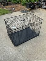 Medium wire dog crate placed outdoors on concrete ground, showing front and side view with a black plastic removable base tray and a handle on the top.