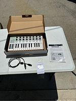 Midi controller Arturia Minilab in box with included USB cable and manual laid out on white table outdoors.