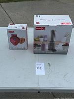 Two boxed kitchen appliances on white table outside: Dash Mini Waffle Maker and Dash Arctic Chill Blender.