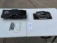 Two model cars displayed on a white table: a black Lego car with exposed engine components and a black Nissan GT-R model on a display base, along with a small bag of Lego parts.