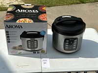 Aroma multicooker and original box shown outdoors on a table, stainless steel finish with black lid and digital buttons on front.