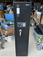 Full front view of tall black safe in a cluttered room showing the handle, keypad, and general condition.