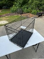 Small black wire dog crate shown on a white folding table outdoors, showcasing the wire structure and plastic tray base.