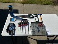Full view of all tools on a white table outside, inclusive of gloves, saw, hammer, screwdrivers, drill bit set, and a metal tool.