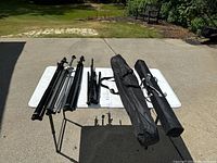 View showing four tripods and two carrying bags with extension poles, displayed on a white table on a driveway outdoors.