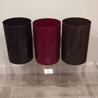 Three cylindrical vintage plastic waste baskets in black, burgundy, and brown shown front view on clear acrylic display stand.