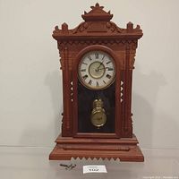 Front view of antique mantle clock showing ornate woodwork, Roman numeral clock face, pendulum, and winding key.