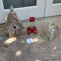 Photo of three items including wooden bird house, red hummingbird feeder, and clear glass bird feeder globe on concrete