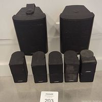 Front view of two larger Audio Source outdoor speakers behind five smaller Bose Jewel Cube speakers, showing wear and Bose logos