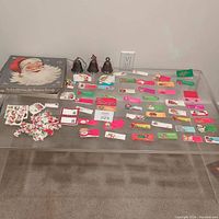 Overview of three Kirk Stieff silver plated bell ornaments and an assortment of vintage Christmas gift tags, stickers, and wrapping paper laid out on a glass table with a Santa Claus box.
