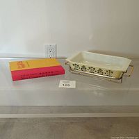 Photo displaying the Pyrex casserole dish, metal stand, and vintage cookbook together on a clear surface with auction lot card number 165.