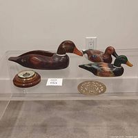 Three painted wooden duck decoys of varying sizes, German wood barometer with round wooden base, and decorative metal trivet on clear acrylic table against white background.