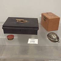 Photo showing four items: a black rectangular metal box with brass handle, a small wooden box with brass acorn-shaped latch, a small brass box shaped like an acorn, and a small round cloisonné brass trinket box with butterflies.