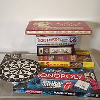 Photo showing stack of board games including By Jove, Ticket to Ride Europe, Dread Pirate, Class Struggle, Snakes & Ladders, Monopoly Rolling Stones, and a 12" dart board plus Master Mind game.