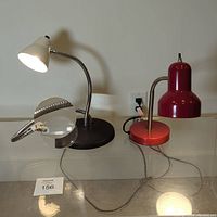 View of the two table lamps and two magnifiers as a lot. Combination lamp shows white conical lamp and double magnifying lens on flexible metal arms. Red lamp with curved arm and round base beside it.