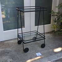 Front view of the vintage metal and glass rolling bar cart on a concrete floor outside, showing two shelves and wheels.