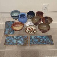 Photo showing 8 pottery items including bowls and pots arranged around a small card with lot number, along with 3 rectangular ceramic tiles with blue and brown mottled glaze.