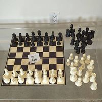 Two full weighted chess sets displayed with black and white pieces arranged on and beside a cardboard chess board. Pieces include extra queens. No chips or cracks visible.