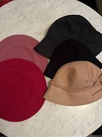 Five wool hats on a white marble surface including three berets (red, dark pink, light pink) and two bucket-style hats (black striped, light brown with braided band and bow)