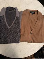 Two men's wool sweaters, one gray V-neck with diamond pattern and one light brown cardigan with buttons.