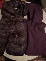 Photo showing the dark brown puffer jacket and purple hooded jacket laid side by side to show fabrics and colors.