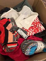 Box of assorted new clothing items piled together showing variety of colors, patterns, and fabrics with tags attached.