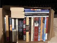 Top-down view of the assortment of books in a cardboard box, showing spines and partial covers with titles and author names.