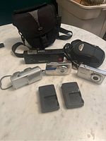 Four cameras, two chargers, and two camera bags arranged on a table, showing assortment of items in lot.