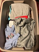 Photo showing a plastic bin filled with assorted children's clothing items including gray tops and a blue onesie with Minnie Mouse print.