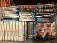 Overhead view of a collection of Blu-ray and DVD movies arranged in rows on a patterned rug. Includes multiple copies or volumes of anime titles, documentary series, and assorted movies in various plastic cases.