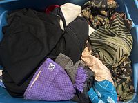 Overview of assorted clothing items in blue bin showing black clothing, patterned scarves, purple knitted hat and blue gloves.