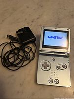 Game Boy Advance SP handheld console powered on showing GAME BOY logo, with charger cable and adapter.